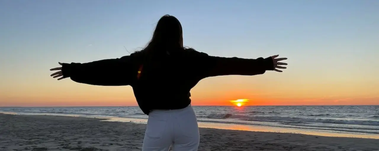 Eine Person steht mit ausgebreiteten Armen am Strand und schaut auf den Sonnenuntergang über dem Meer